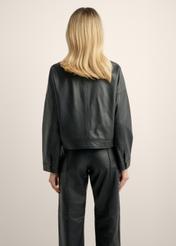 Жіноча коротка шкіряна куртка чорного кольору KURDS-0629-99(Z26), вигляд виробу ззаду на моделі. Видно половину силуету.